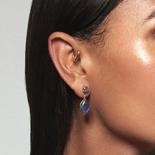 A model wearing Juraster’s 9ct Gold Blue Labradorite Earring Charm, Adventure, crafted from Fairmined yellow gold and hand-cut blue labradorites attached to a matching earring stud and a yellow gold hoop in her forward helix.