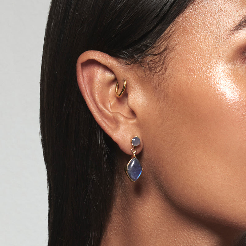 A model wearing Juraster’s 9ct Gold Blue Labradorite Earring Charm, Adventure, crafted from Fairmined yellow gold and hand-cut blue labradorites attached to a matching earring stud and a yellow gold hoop in her forward helix.