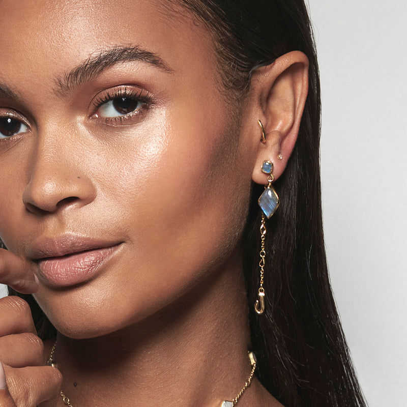 A model wearing Juraster’s 9ct Gold Blue Labradorite Stud Earring, Adventure, crafted from Fairmined yellow gold and hand-cut blue labradorites and matching earring charm. She also wears a yellow gold hoop and diamond stud with a yellow gold dangle earring charm.