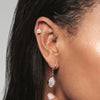 A model wearing Juraster’s 9ct Gold Rainbow Moonstone Stud Earring, Adventure, crafted from Fairmined white gold and hand-cut rainbow moonstone, in her cartilage. She also wears two sterling silver hoops, one in her forward helix and one in her lobe with a sterling silver rose quartz earring charm.