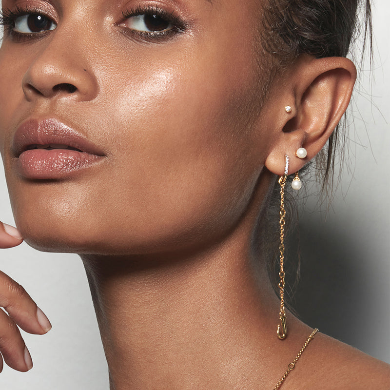 A model wearing Juraster’s Akoya Pearl Stud Earring, Beachcomber, crafted from Fairmined yellow gold and a responsibly-farmed pearl with a matching earring charm. She is also wearing two yellow gold diamond earrings, one a stud and one pavé diamond hoop with a yellow gold dangle earring charm.