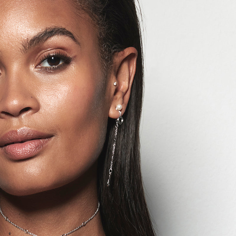 A model wearing Juraster’s Sterling Silver Dangle Earring Charm, Discovery, crafted from Fairmined sterling silver, alongside a sterling silver diamond-shaped earring charm, attached to a sterling silver pearl earring stud. She also wears a white gold diamond stud in her tragus.