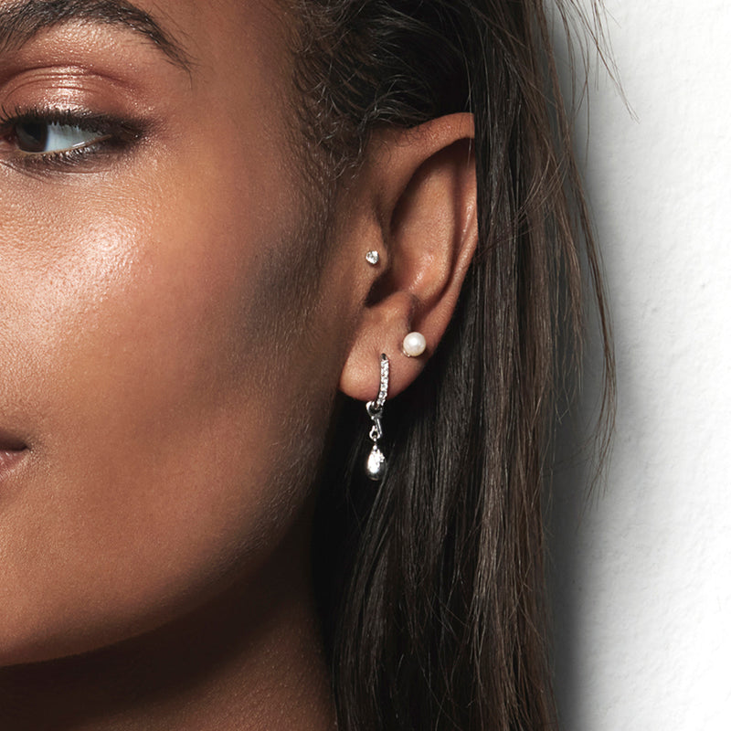 A model wearing Juraster’s 9ct Gold Classic Diamond Stud Earring, Light-Ray, crafted from Fairmined white gold and conflict-free diamonds. She also wears a sterling silver pearl stud in her second and a white gold pavé diamond hoop in her lobe with a sterling silver star droplet earring charm.