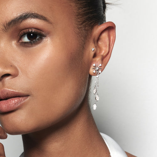 A model wearing Juraster’s 9ct White Gold Diamond Earring Jacket, Snowflake, crafted from Fairmined white gold and conflict-free diamonds, and a white gold diamond tassel earring charm, attached to a white gold diamond stud earring. She also wears a white gold diamond stud earring in her tragus.